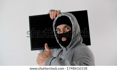 Thief in black balaclava pretty showing thumb. Offender with a closed face. Riots, robbery and marauding.Theft of technology. Man carries a TV on his shoulder. 