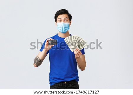 Money, lifestyle, insurance and investment concept. Excited good-looking young male student making deposit on banking account, showing credit card and cash dollars, stand in medical mask