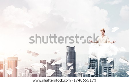 Man in white clothing keeping eyes closed and looking concentrated while meditating on cloud among flying paper planes with cityscape view on background.