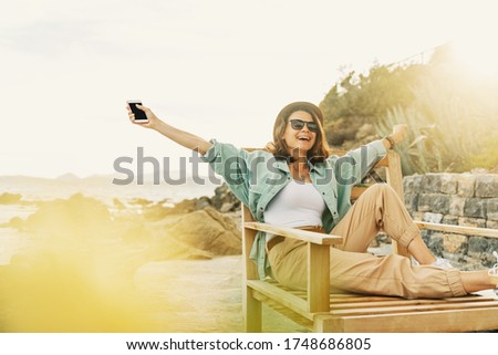 Outdoor summer smiling lifestyle portrait of pretty young woman having fun with smartphone in hands. Making pictures in hipster style glasses and hat