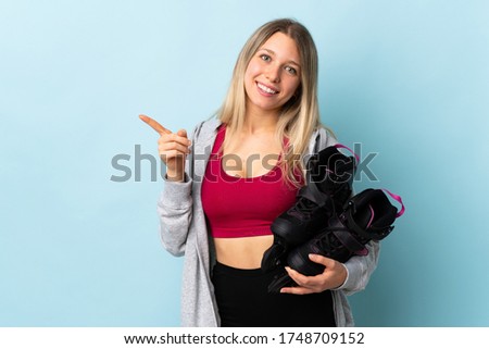 Young blonde woman holding a roller skates isolated on pink background pointing finger to the side