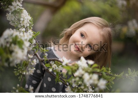 very beautiful baby girl with blond hair on a background of a flowering cherry tree. large portrait. girl smiles and looks away. on a child a gray dress in large white peas. image with selective focus