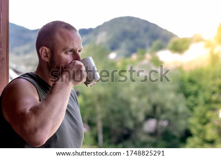 Photo of smiling young man standing on the balcony with cup of coffee.Handsome man having his morning coffee on a balcony and daydreaming.Caucasian man looking away and sipping coffee in white cup.