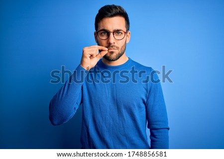 Young handsome man with beard wearing casual sweater and glasses over blue background mouth and lips shut as zip with fingers. Secret and silent, taboo talking Royalty-Free Stock Photo #1748856581