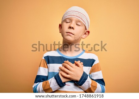 Young little caucasian kid injured wearing medical bandage on head over yellow background smiling with hands on chest with closed eyes and grateful gesture on face. Health concept.