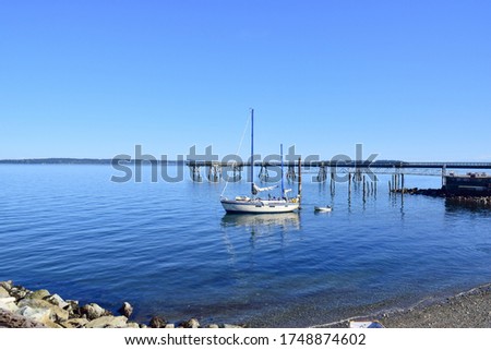 White fishing boat floating in blue sea with its reflection, there is a rock beach foreground and bridge with a smooth gradient clear blue sky with no cloud background for copy space on top of image