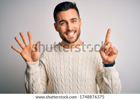 Young handsome man wearing casual sweater standing over isolated white background showing and pointing up with fingers number six while smiling confident and happy.