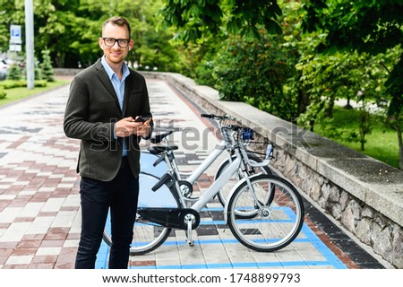 A young businessman in smart casual using app for rent city bike. A guy wearing glasses is typing on the smartphone, parking of city bicycles on the background