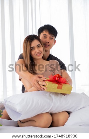 The couple lovers hold a golden gift box  in thier hands on the bed in the bedroom decorated with red heart balloons on Valentine's Day.