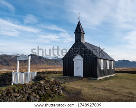 Small black church Búðakirkja with white doors and windows and gate and stone wall along with view of mountains covered by snow on peninsula Snaefellsness during sunny autumn day