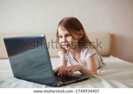 Close-up portrait of a baby girl with a laptop on the bed. The concept of digital communication, digital addiction, entertainment, distance learning online, home.