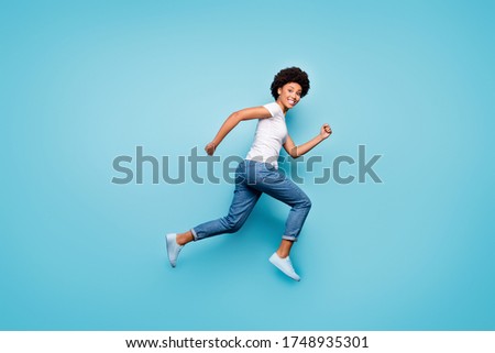 Full body photo of funky dark skin curly lady jumping high rushing marathon finish line coming first active sporty person wear casual white t-shirt jeans isolated blue color background