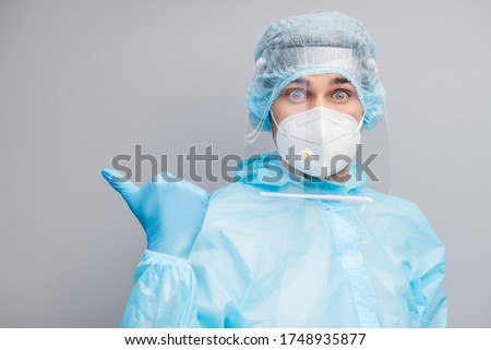 Closeup photo of shocked guy doc virologist direct thumb finger empty space show free vaccination center wear mask hazmat blue uniform suit protective goggles isolated grey color background