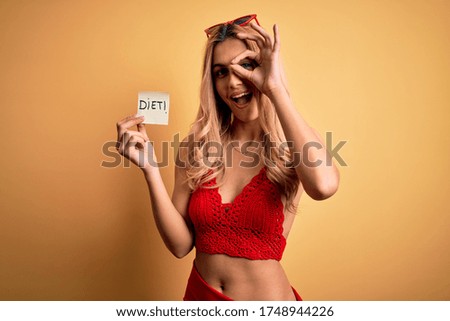 Beautiful blonde slim woman on vacation wearing bikini holding reminder with diet message with happy face smiling doing ok sign with hand on eye looking through fingers