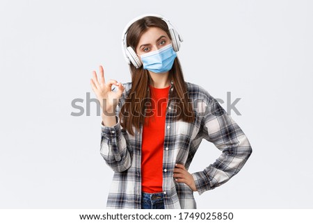 Social distancing, leisure and lifestyle on covid-19 outbreak, coronavirus concept. Pleased good-looking woman in medical mask and headphones, listening music, show okay sign, approve or like