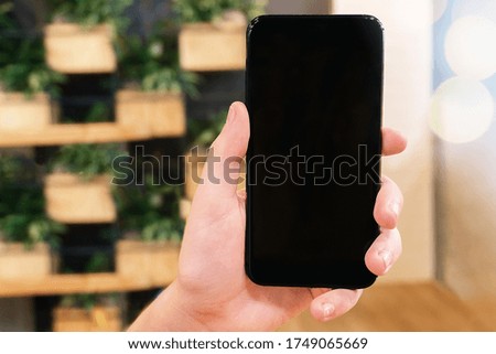 Close up male hand holding black mobile phone on background of wooden vases with live green flowers. World mobile shopping is in smartphone. Concept of e-commerce, focus on hand.