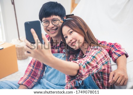 Asian couple husband and wife taking selfie, smiling woman holding phone, happy man posing for photo with girlfriend, family having fun together with gadget in new room.