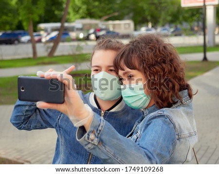 two women, a mother and daughter, in blue denim jackets and medical masks, take selfies with a smartphone on the street during the coronavirus pandemic
