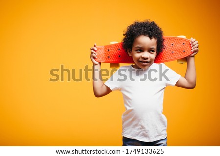 Black boy child in a white T-shirt holds a skate on his shoulders on an orange background in the studio.