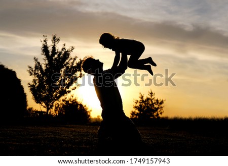 A silhouette of a happy father lovingly lifting his little toddler child over his head and playing outside at sunset.