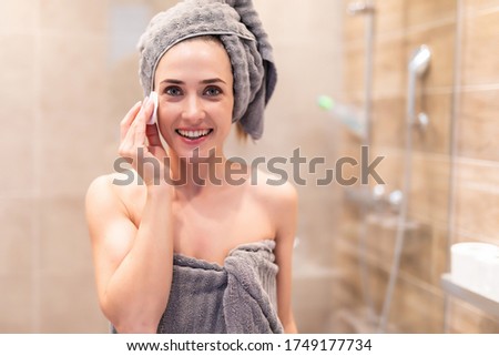 Head shot close up beautiful woman wearing white bath towel on head cleaning skin with cotton pad after shower, pretty female with healthy skin standing in bathroom, looking at camera