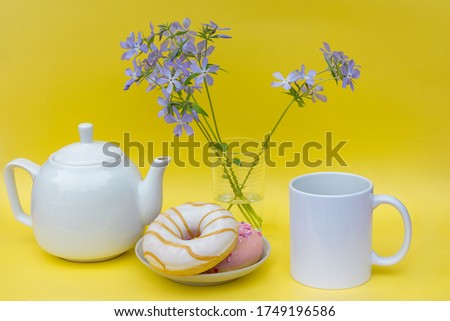 On a yellow background there is a white kettle and a mug, without logos. On the plate are beautiful, freshly baked doughnuts. In the vase there are delicate blue chicks. Romantic breakfast