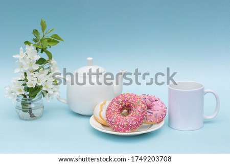 On a blue background, a white teapot and a mug without logos on a plate are donuts, flowers in a vase. In theory, this is a beautiful, delicate, festive, romantic breakfast. Photo in pastel colors.