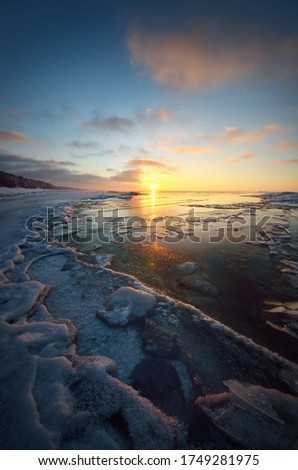 Panoramic view of the frozen Baltic sea shore at sunset. Ice fragments close-up, snow-covered coniferous forest in the background. Colorful cloudscape. Symmetry reflections on the water. Latvia