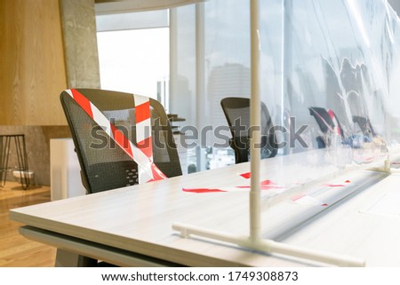 Office chairs marked with white and red tape looking through the plastic physical wall. New normal Concept for social distancing in offices during the Covid-19 pandemic. Precaution and safe workspace.