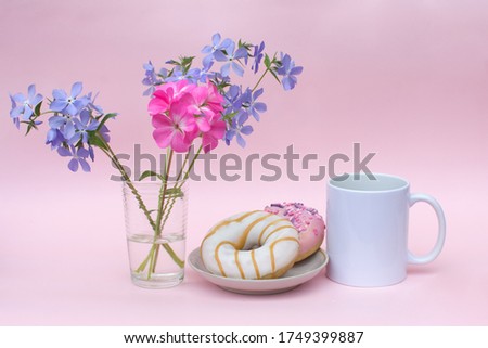 On a pink background-a white mug without logos. There are 2 delicious doughnuts on the plate. There are blue and pink flowers in the vase. Photos in pastel colors. Advertising a delicious Breakfast.