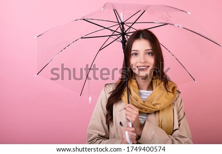 Portrait of beautiful young brunette lady wearing an autumn coat and scarf standing, smiling tenderly, isolated on a pink background, holding an umbrella, hiding from the rain, bad weather.Copy space.