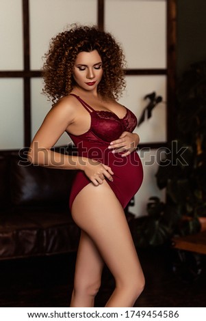 Portrait of curly pregnant european woman in a red swimsuit, happy stylish pregnant woman in dark interior, happiness of motherhood, future mother, waiting for a child