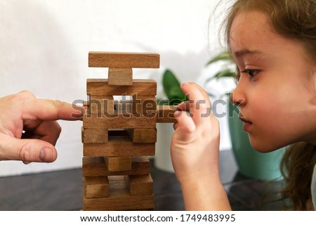 Father and daughter playing game tumble tower from wooden block together. Man's hand pushes block of wood, and girl pulls it out. Man helps child win. Royalty-Free Stock Photo #1749483995