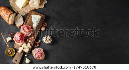 Banner with delicious snack of prosciutto, cheese, nuts and honey. Wooden chopping board. Dark textured background. Appetizer, aperitif, snack table. Top view. Space for text. Royalty-Free Stock Photo #1749518678