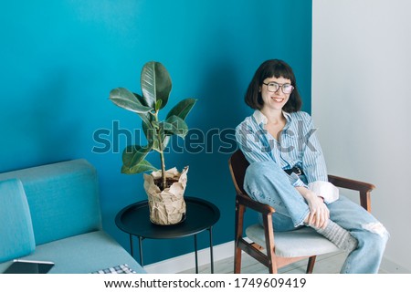 
Cute girl in glasses and a blue shirt works on a laptop while sitting in a vintage armchair at home, drinks coffee from a mug. White-blue background, next to it is a flower in a pot.