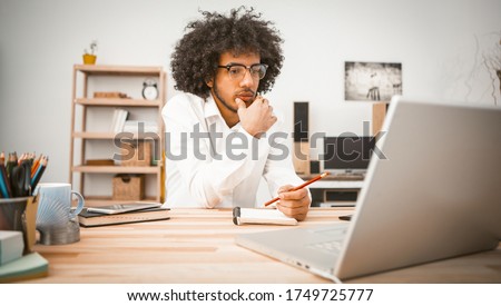 Pensive young man is engaged at home in front of computer. Arab student or businessman taking notes in a notebook. Home education concept. Toned image. Royalty-Free Stock Photo #1749725777
