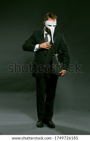 Masked businessman puts large wad of money in his breast pocket holding briefcase under his arm. Man in fancy white masks full-length stands on gray background. Corruption concept. Toned image.