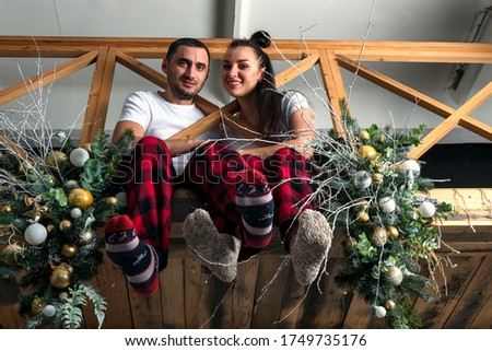 A happy couple in white t-shirts and red and black checked trousers are sitting nearby on a wooden balcony decorated with Christmas decorations.