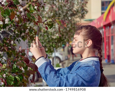 a young girl in a blue jeans jacket on the street takes photos of blossoms on her smartphone for the social networks