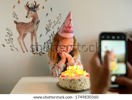 birthday of a little girl, sitting near the table, looking at a cake with candles, in defocus in the foreground a photo is taken on the phone.