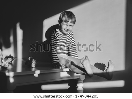 
A little boy in a vest sits on a white table. The child's legs are raised up. On the table behind the boy are newspaper boats. The boy smiles slyly. Image is black and white with selective focus.
