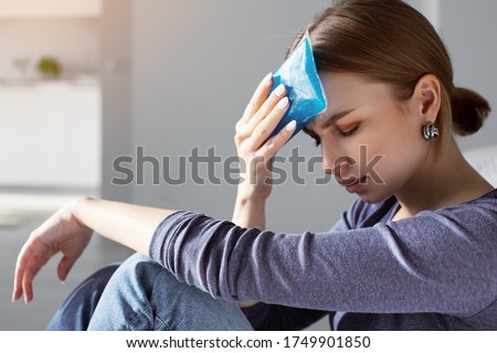 Woman applying ice pack as cold compress on forehead due to headache, migraine, tired after work, sitting indoors in apartment. Put cold on head for relief hurt and fever Royalty-Free Stock Photo #1749901850