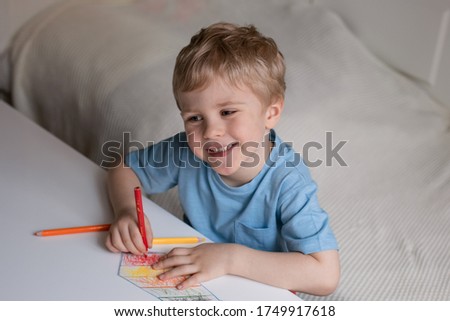 Cute little boy with blond hair and blue T-shirt draws colored pencils at home. Draws at the white table