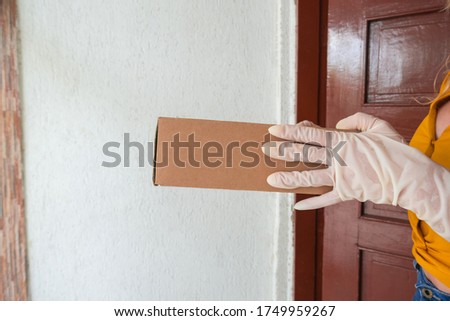 Safe home delivery of packages during the coronavirus pandemic. The hands of a woman wearing white medical gloves hold a cardboard box against a light background