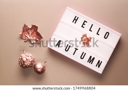 Lightbox with text Hello Autumn. Light bronze color paper background. Flat lay with dry sycamore leaf, bronze metallic pumpkin and small apples.