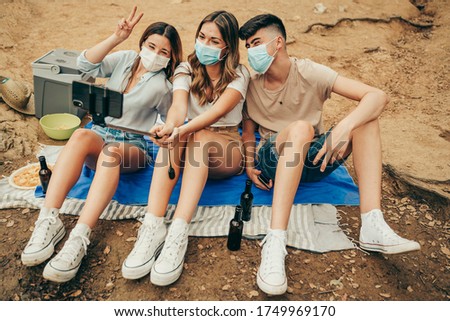Masked young friends taking a photo with a photo stick on a camping trip in the middle of nature during the pandemic.