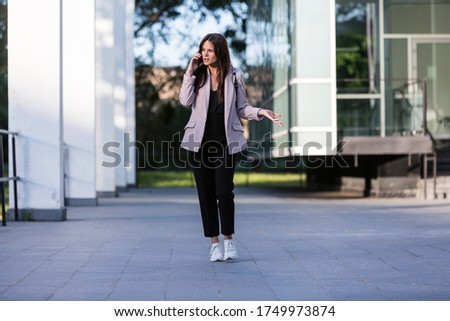 A young beautiful girl is walking while talking on her cell phone. She is smiling, business-trained with modern architecture in the background.