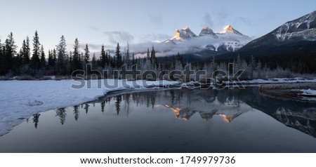 Panoramic winter sunrise shot from Canadian Rockies with mountain reflected in water, shot at Three Sisters Mountain, Canmore, Alberta, Canada Royalty-Free Stock Photo #1749979736