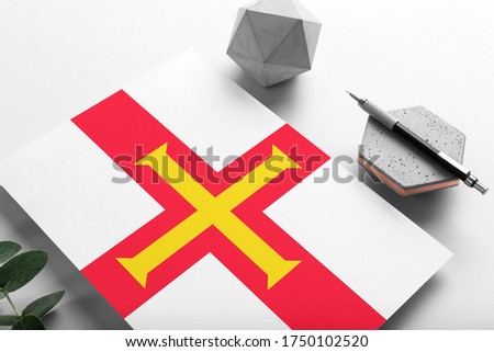 Guernsey flag on minimalist paper background. National invitation letter with stylish pen on stone. Communication concept.