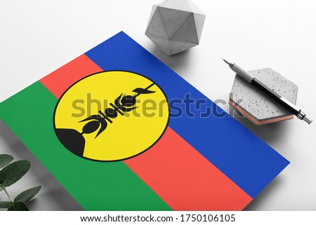 New Caledonia flag on minimalist paper background. National invitation letter with stylish pen on stone. Communication concept.
